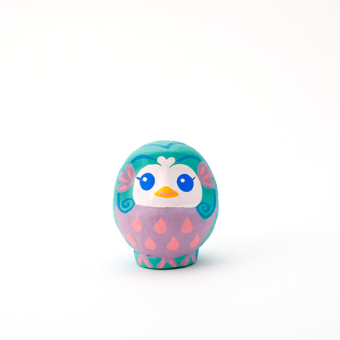 An Imai Daruma Naya Amabie-sama, lucky creature papier-mache Japanese doll, with a purple stomach with pink scales, a turquoise head and white face, featuring blue eyes and yellow beak.