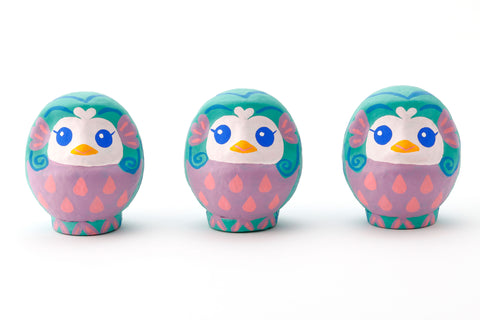 Three Imai Daruma Naya Amabie-sama, lucky creature papier-mache Japanese dolls. Each one has a purple stomach with pink scales, a turquoise head and white face, featuring blue eyes and yellow beak.