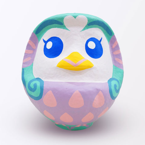 A large Imai Daruma Naya Amabie-sama, lucky creature papier-mache Japanese doll, with a purple stomach with pink scales, a turquoise head and white face, featuring blue eyes and yellow beak.