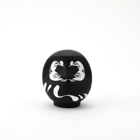 A small black Imai Daruma Naya Designer’s Daruma Japanese papier-mache doll, featuring a pattern of white eyebrows and beard and a three swirl lines across its body.
