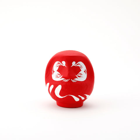 A small red Imai Daruma Naya Designer’s Daruma Japanese papier-mache doll, featuring a pattern of white eyebrows and beard and a three swirl lines across its body.