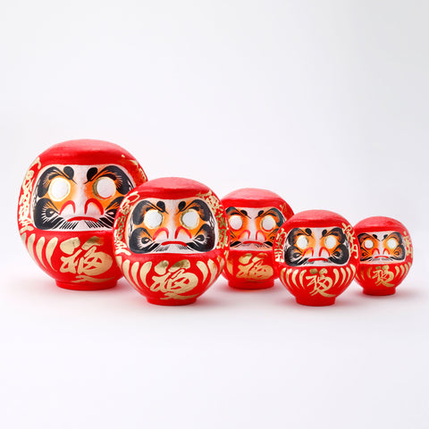 A group of five different sized red Fuku-iri papier-mache daruma dolls, made by Imai Daruma Naya. All feature rotund bodies, decorative patterns for eyebrows and beards and gold body lines and kanji lettering.