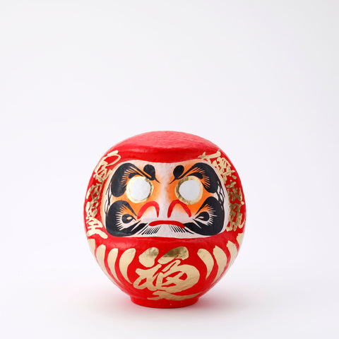 A 20-centimeter tall red Fuku-iri lucky daruma doll, made by Imai Daruma Naya, featuring a rotund body and decorative patterns for eyebrows and beard and gold body lines and kanji lettering.