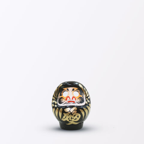 A 9-centimeter tall black Fuku-iri lucky daruma doll, made by Imai Daruma Naya, featuring a rotund body and decorative patterns for eyebrows and beard and gold body lines and kanji lettering.