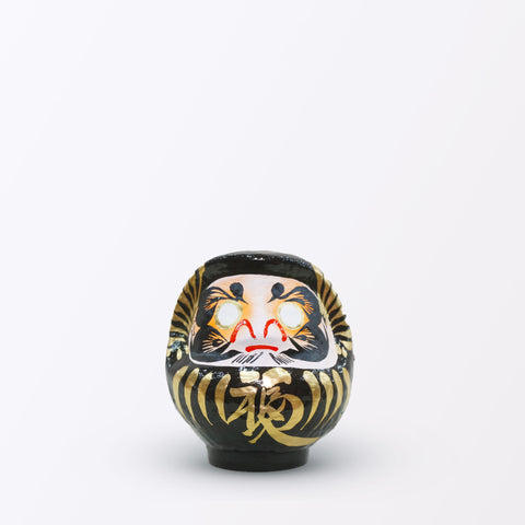 A 12-centimeter tall black Fuku-iri lucky daruma doll, made by Imai Daruma Naya, featuring a rotund body and decorative patterns for eyebrows and beard and gold body lines and kanji lettering.