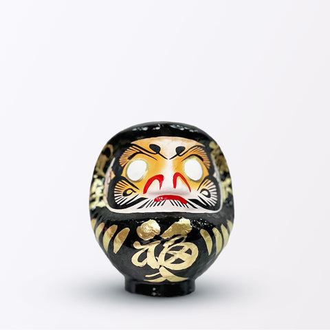 A 15-centimeter tall black Fuku-iri lucky daruma doll, made by Imai Daruma Naya, featuring a rotund body and decorative patterns for eyebrows and beard and gold body lines and kanji lettering.