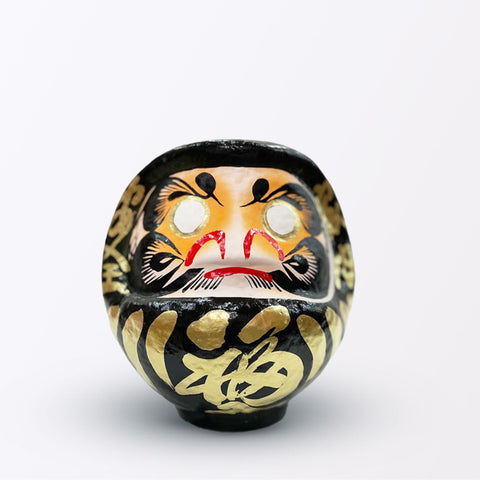 A 17-centimeter tall black Fuku-iri lucky daruma doll, made by Imai Daruma Naya, featuring a rotund body and decorative patterns for eyebrows and beard and gold body lines and kanji lettering.