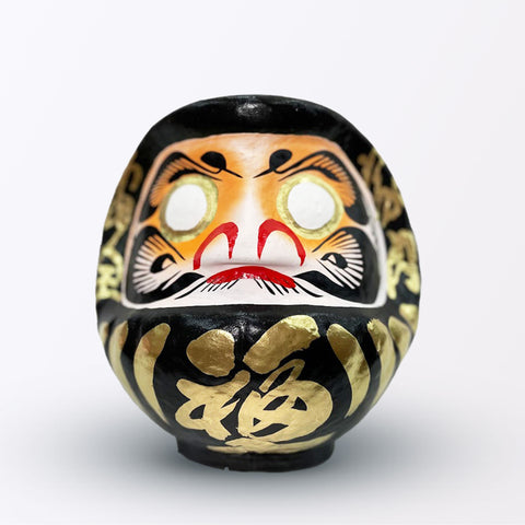 A 20-centimeter tall black Fuku-iri lucky daruma doll, made by Imai Daruma Naya, featuring a rotund body and decorative patterns for eyebrows and beard and gold body lines and kanji lettering.
