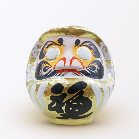 A 20-centimeter tall gold Fuku-iri lucky daruma doll, made by Imai Daruma Naya, featuring a rotund body and decorative patterns for eyebrows and beard and white body lines with black kanji lettering.
