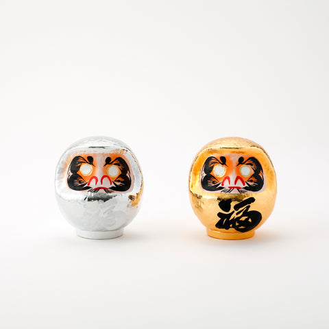 A pair of shiny Platinum Daruma lucky dolls — one silver and one gold — made by Imai Daruma Naya. Both feature rotund bodies and decorative patterns for eyebrows and beards. The silver has kanji lettering in white, the gold has lettering in black.