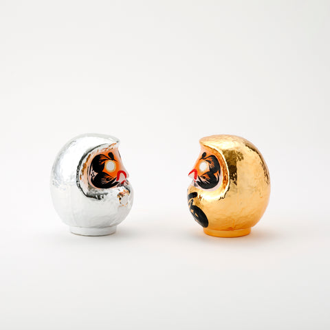 A pair of facing shiny Platinum Daruma lucky dolls — one silver and one gold — made by Imai Daruma Naya. Both feature rotund bodies and decorative patterns for eyebrows and beards. The silver has kanji lettering in white, the gold has lettering in black.