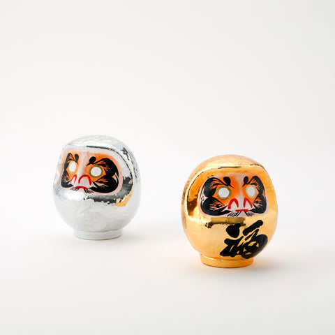 A pair of shiny Platinum Daruma lucky dolls — one silver and one gold — made by Imai Daruma Naya. Both feature rotund bodies and decorative patterns for eyebrows and beards. The silver has kanji lettering in white, the gold has lettering in black.