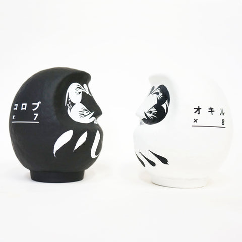 Two facing Imai Daruma Naya Designer’s Daruma Japanese papier-mache dolls — one black (left) and one white (right), each featuring a decorative pattern of eyebrows and beard and a three swirl lines in white across the body. The black features Japanese katakana for “korobu x 7” meaning to fall seven times, while the white features “Okiru x 8,” meaning to get up 8 times.