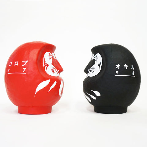 Two facing Imai Daruma Naya Designer’s Daruma Japanese papier-mache dolls — one red (left) and one black(right), each featuring a decorative pattern of eyebrows and beard and a three swirl lines in white across the body. The red features Japanese katakana for “korobu x 7” meaning to fall seven times, while the black features “Okiru x 8,” meaning to get up 8 times. 