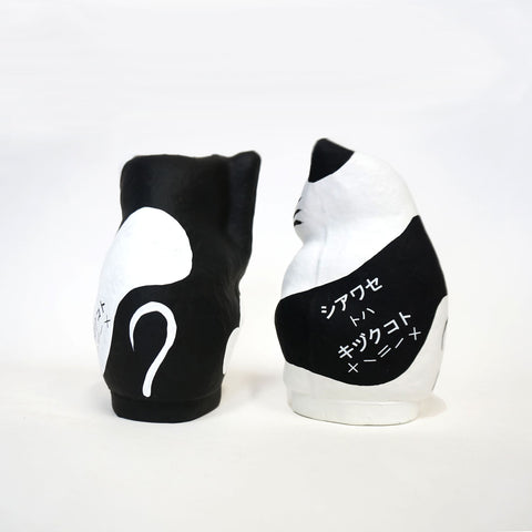 A pair of Imai Daruma Naya Designer Neko cat Japanese manekineko papier-mache dolls shown from the rear — one white and one black — both showing the phrase "Happiness is a gift" in katakana..