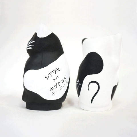 A pair of Imai Daruma Naya Designer Neko cat Japanese manekineko papier-mache dolls shown from the rear — one white and one black — both showing the phrase "Happiness is a gift" in katakana..