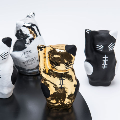 A gold Imai Daruma Naya Platinum Designer Neko cat Japanese manekineko papier-mache doll standing with similar white, silver and black versions, featuring white patches and whiskers and raising its right paw to beckon good luck.