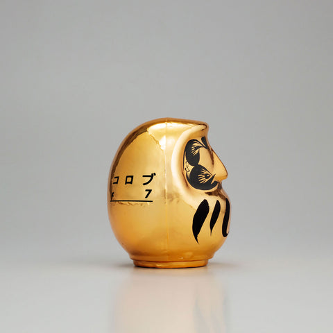 A side view of a shiny, gold Platinum Designer’s Daruma lucky doll, made by Imai Daruma Naya. The rotund doll features decorative patterns for its eyebrows and beard, and black curvy lines across its body.