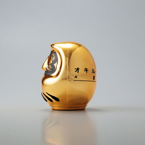 A side view of a shiny, gold Platinum Designer’s Daruma lucky doll, made by Imai Daruma Naya. The rotund doll features decorative patterns for its eyebrows and beard, and black curvy lines across its body.