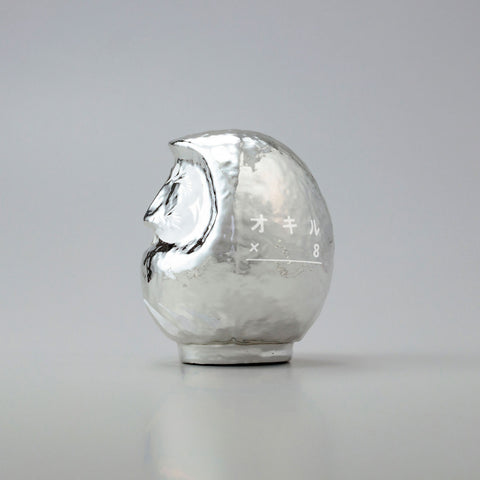 A side view of a shiny, silver Platinum Designer’s Daruma lucky doll, made by Imai Daruma Naya. The rotund doll features decorative patterns for its eyebrows and beard, and white curvy lines across its body.