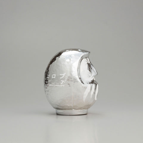 A side view of a shiny, silver Platinum Designer’s Daruma lucky doll, made by Imai Daruma Naya. The rotund doll features decorative patterns for its eyebrows and beard, and white curvy lines across its body.