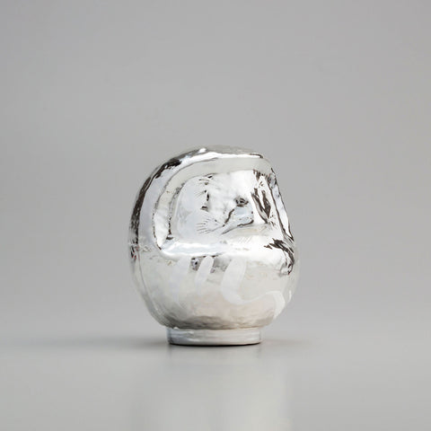 A shiny, silver Platinum Designer’s Daruma lucky doll, made by Imai Daruma Naya. The rotund doll features decorative patterns for its eyebrows and beard, and white curvy lines across its body.