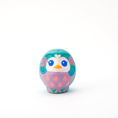 An Imai Daruma Naya Amabie-sama, lucky creature papier-mache Japanese doll, with a purple stomach with pink scales, a turquoise head and white face, featuring blue eyes and yellow beak. 