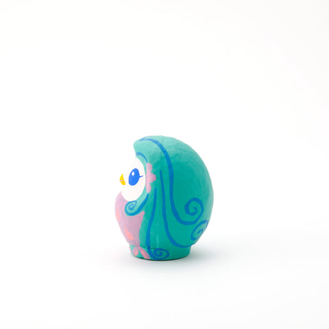 A side view of an Imai Daruma Naya Amabie-sama, lucky creature papier-mache Japanese doll, with a purple stomach with pink scales, a turquoise head and white face, featuring blue eyes and yellow beak. 