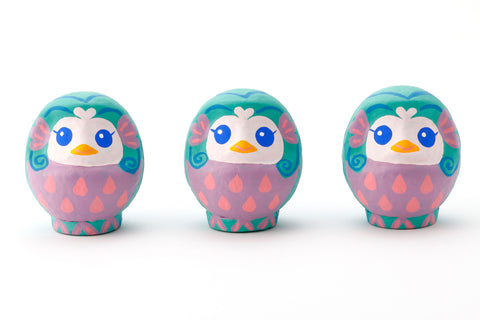 Three Imai Daruma Naya Amabie-sama, lucky creature papier-mache Japanese dolls. Each one has a purple stomach with pink scales, a turquoise head and white face, featuring blue eyes and yellow beak. 