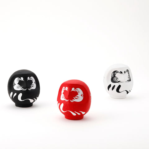 Three Imai Daruma Naya Designer’s Daruma Japanese papier-mache dolls — one black, one red and one white, each featuring a decorative pattern of eyebrows and beard and a three swirl lines across its body.
