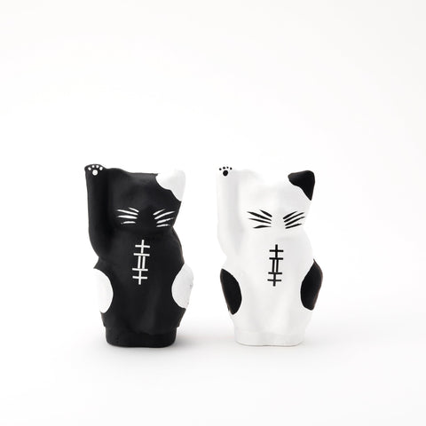 A pair of Imai Daruma Naya Designer Neko cat Japanese manekineko papier-mache dolls — one white and one black — both raising their right paws to beckon good luck. The black features white patches and whiskers, the white has black patches and whiskers.