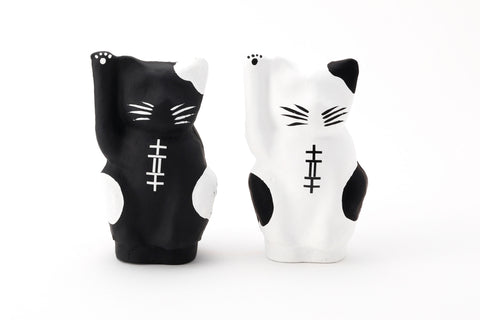  A pair of Imai Daruma Naya Designer Neko cat Japanese manekineko papier-mache dolls — one white and one black — both raising their right paws to beckon good luck. The black features white patches and whiskers, the white has black patches and whiskers.