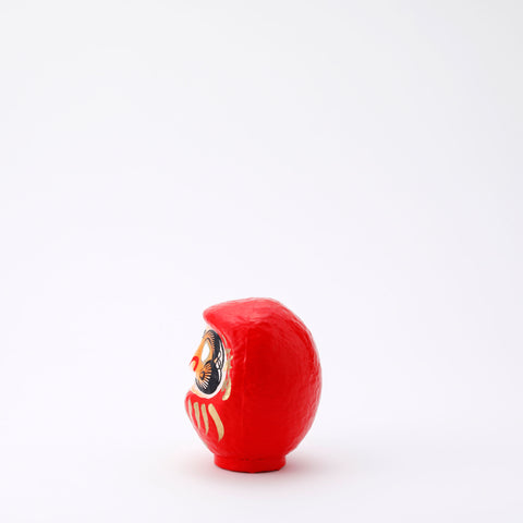 A side view of a 9-centimeter tall red Fuku-iri lucky daruma doll, made by Imai Daruma Naya, featuring a rotund body and decorative patterns for eyebrows and beard and gold body lines and kanji lettering.