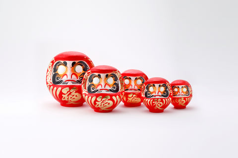 A group of five different sized red Fuku-iri papier-mache daruma dolls, made by Imai Daruma Naya. All feature rotund bodies, decorative patterns for eyebrows and beards and gold body lines and kanji lettering.
