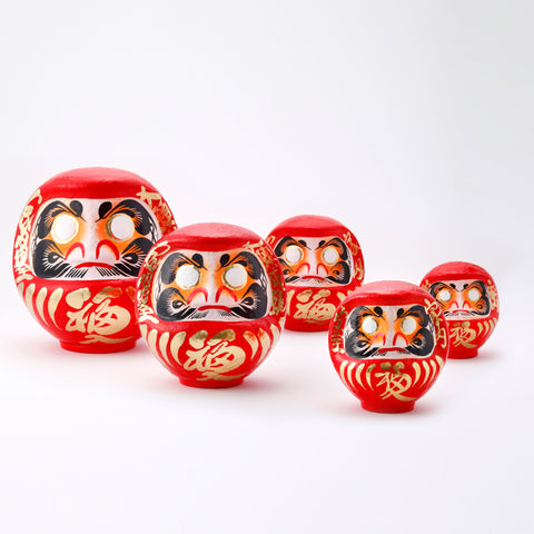 A group of five different sized red Fuku-iri papier-mache daruma dolls, made by Imai Daruma Naya. All feature rotund bodies, decorative patterns for eyebrows and beards and gold body lines and kanji lettering.
