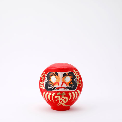 A 12-centimeter tall red Fuku-iri lucky daruma doll, made by Imai Daruma Naya, featuring a rotund body and decorative patterns for eyebrows and beard and gold body lines and kanji lettering.