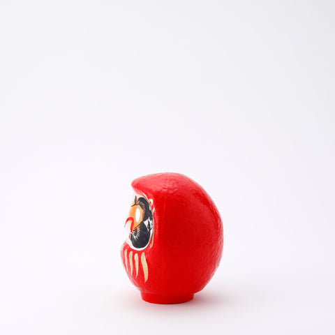 A side view of a 12-centimeter tall red Fuku-iri lucky daruma doll, made by Imai Daruma Naya, featuring a rotund body and decorative patterns for eyebrows and beard and gold body lines and kanji lettering.