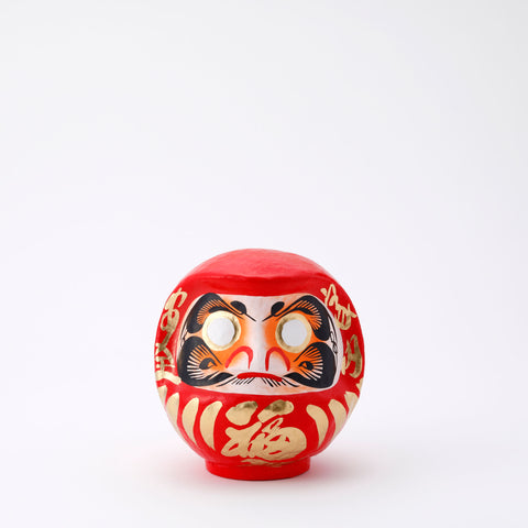 A 15-centimeter tall red Fuku-iri lucky daruma doll, made by Imai Daruma Naya, featuring a rotund body and decorative patterns for eyebrows and beard and gold body lines and kanji lettering.