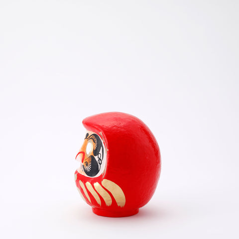 A side view of a 17-centimeter tall red Fuku-iri lucky daruma doll, made by Imai Daruma Naya, featuring a rotund body and decorative patterns for eyebrows and beard and gold body lines and kanji lettering.