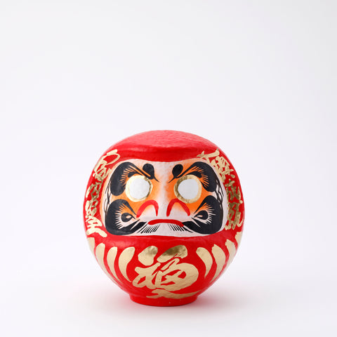  A 20-centimeter tall red Fuku-iri lucky daruma doll, made by Imai Daruma Naya, featuring a rotund body and decorative patterns for eyebrows and beard and gold body lines and kanji lettering.