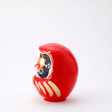 A side view of a 20-centimeter tall red Fuku-iri lucky daruma doll, made by Imai Daruma Naya, featuring a rotund body and decorative patterns for eyebrows and beard and gold body lines and kanji lettering.