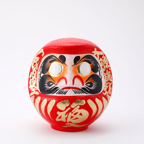 A 23-centimeter tall red Fuku-iri lucky daruma doll, made by Imai Daruma Naya, featuring a rotund body and decorative patterns for eyebrows and beard and gold body lines and kanji lettering.