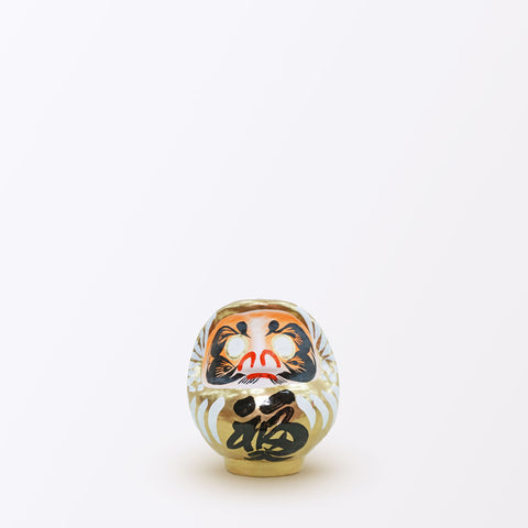 A 9-centimeter tall gold Fuku-iri lucky daruma doll, made by Imai Daruma Naya, featuring a rotund body and decorative patterns for eyebrows and beard and white body lines with black kanji lettering.