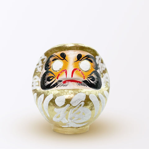 A 17-centimeter tall gold Fuku-iri lucky daruma doll, made by Imai Daruma Naya, featuring a rotund body and decorative patterns for eyebrows and beard and white body lines with black kanji lettering.