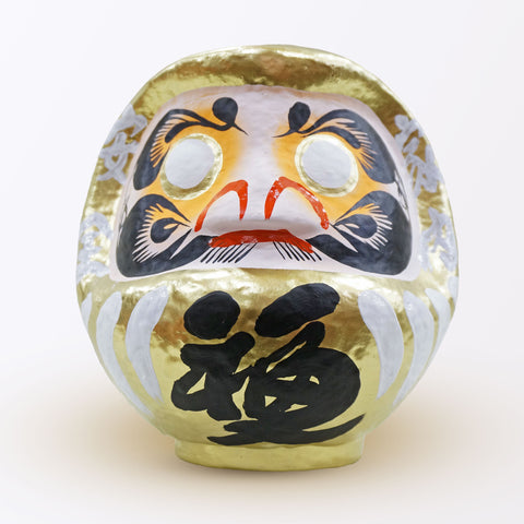 A 23-centimeter tall gold Fuku-iri lucky daruma doll, made by Imai Daruma Naya, featuring a rotund body and decorative patterns for eyebrows and beard and white body lines with black kanji lettering.