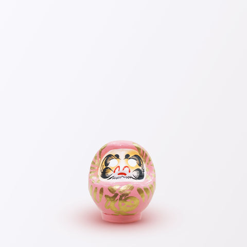 A 9-centimeter tall peach pink Fuku-iri lucky daruma doll, made by Imai Daruma Naya, featuring a rotund body and decorative patterns for eyebrows and beard and gold body lines and kanji lettering.