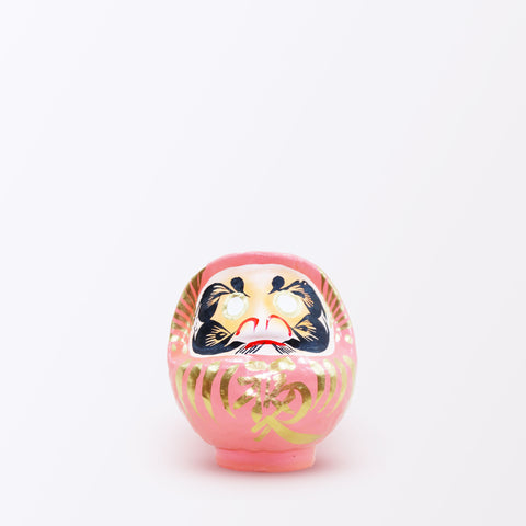 A 12-centimeter tall peach pink Fuku-iri lucky daruma doll, made by Imai Daruma Naya, featuring a rotund body and decorative patterns for eyebrows and beard and gold body lines and kanji lettering.