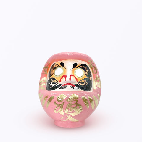 A 15-centimeter tall peach pink Fuku-iri lucky daruma doll, made by Imai Daruma Naya, featuring a rotund body and decorative patterns for eyebrows and beard and gold body lines and kanji lettering.