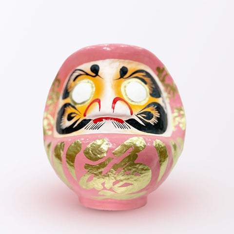 A 20-centimeter tall peach pink Fuku-iri lucky daruma doll, made by Imai Daruma Naya, featuring a rotund body and decorative patterns for eyebrows and beard and gold body lines and kanji lettering.