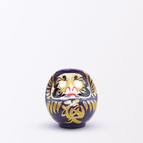 A 12-centimeter tall purple Fuku-iri lucky daruma doll, made by Imai Daruma Naya, featuring a rotund body and decorative patterns for eyebrows and beard and gold body lines and kanji lettering.